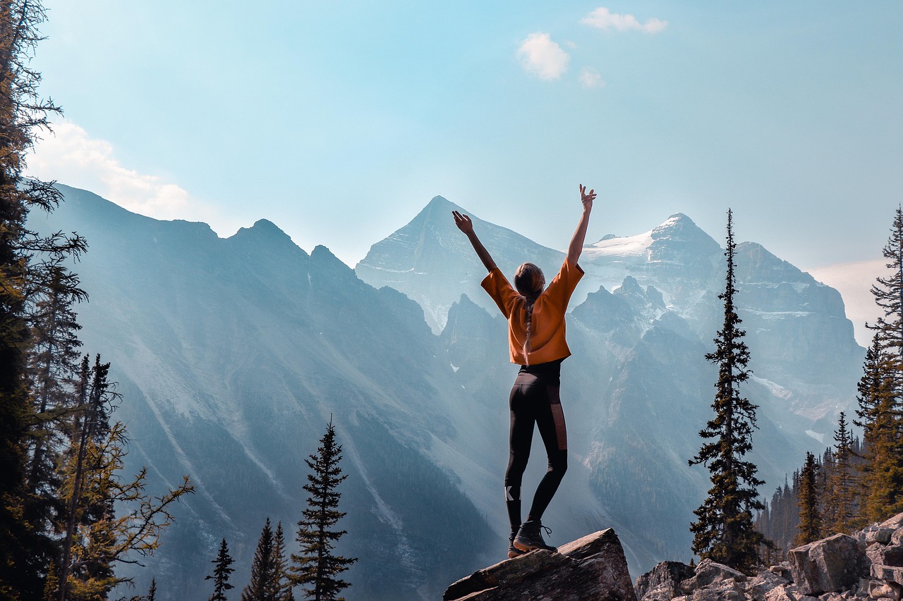 Person with arms raised on a rock overlooking a vast mountain range and pine trees under a blue sky.
