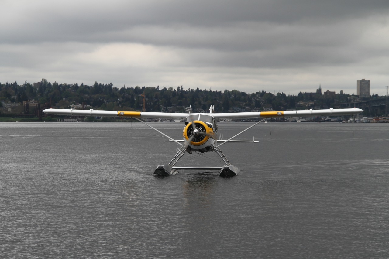 White and yellow floatplane on water under a cloudy sky with trees and buildings in the background.