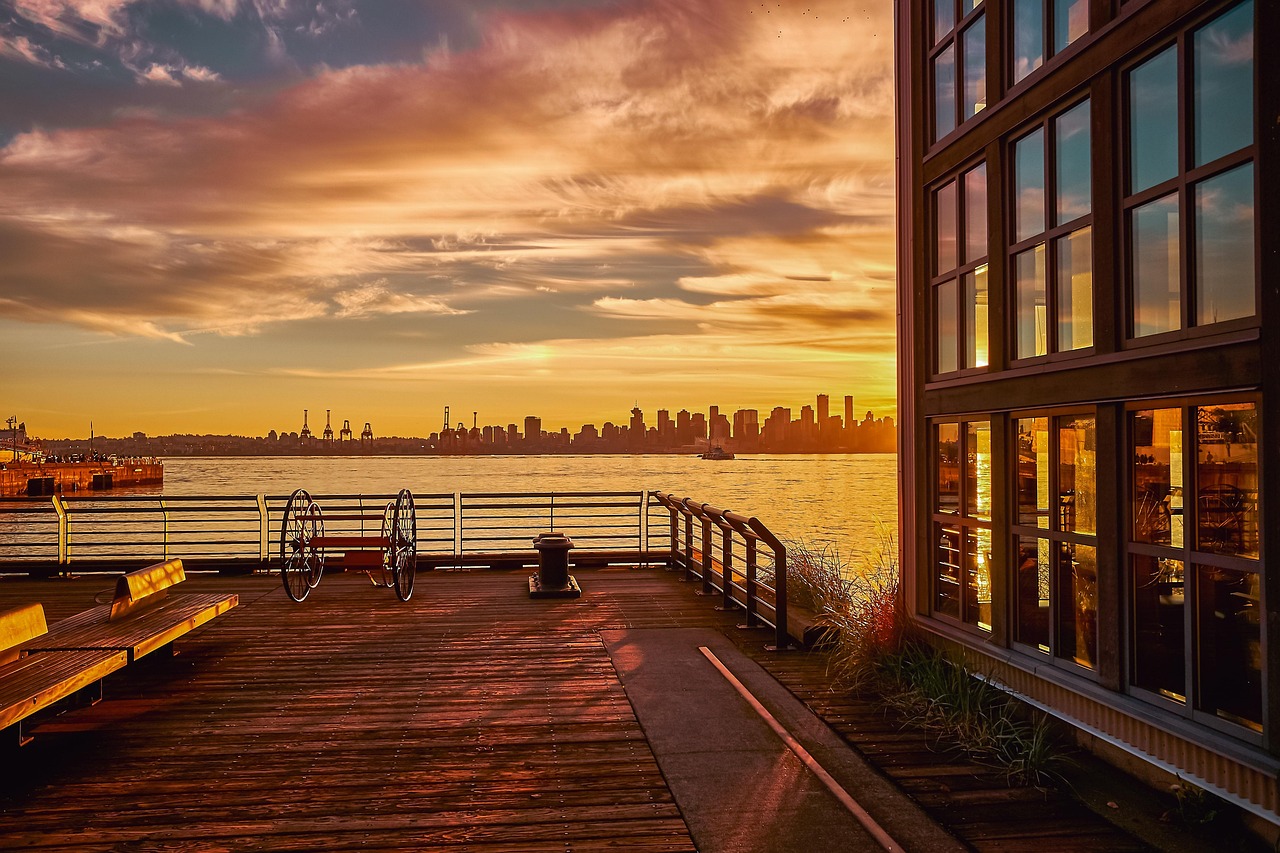 Waterfront promenade at sunset with a city skyline across the water and a modern building on the right.