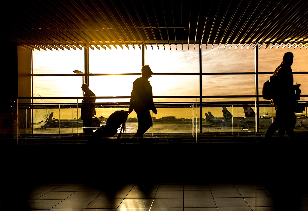 Silhouettes of travelers walk past large windows overlooking an airport tarmac at sunset.