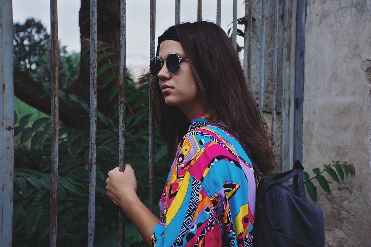 Young person in sunglasses and a colorful shirt holding a metal gate, with a backpack and foliage in the background.