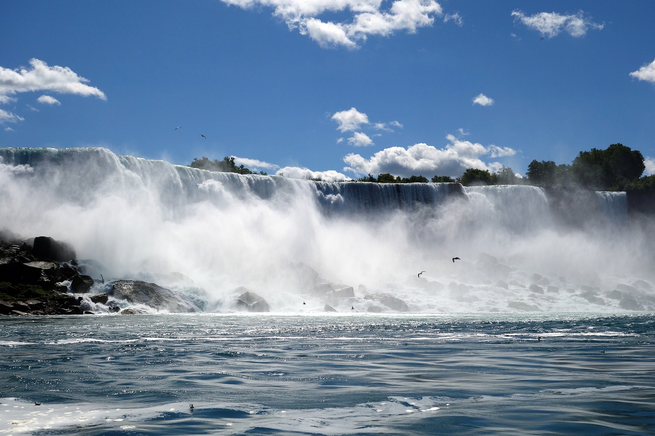 A wide, powerful waterfall cascades into choppy water under a blue sky with white clouds.